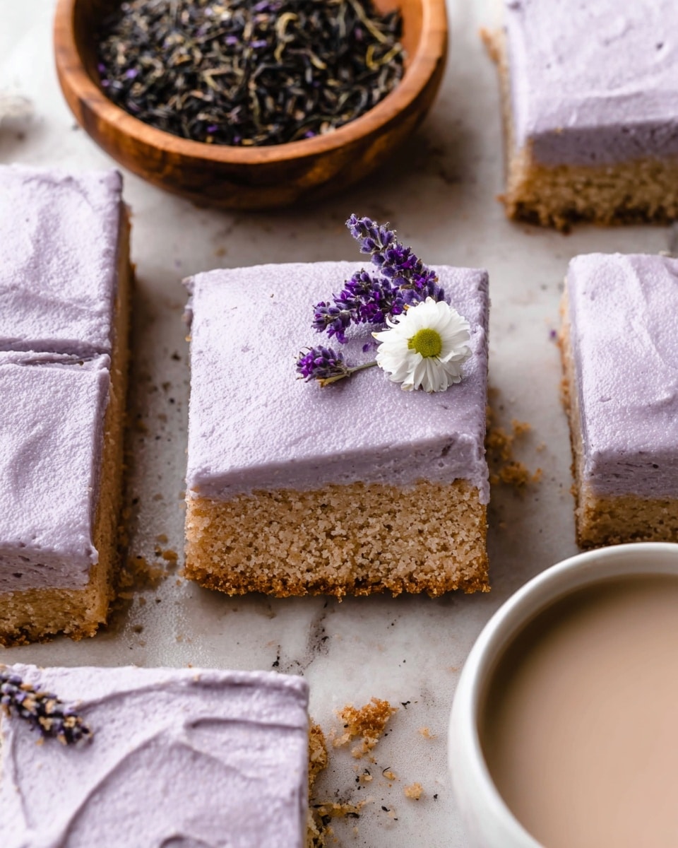 A close-up of square cake pieces with two layers: the bottom layer is light brown and crumbly, and the top layer is thick, smooth, and lavender purple frosting that covers the cake completely. One piece is lifted slightly, showing the cake's texture. One cake piece includes a small white flower with a yellow center and a few small purple flowers on top. The cakes are placed on a white marbled texture surface. To the left, there is a wooden bowl filled with dried lavender flowers and black tea leaves. To the right, part of a white cup filled with a frothy light cream-colored drink is visible. Photo taken with an iphone --ar 4:5 --v 7