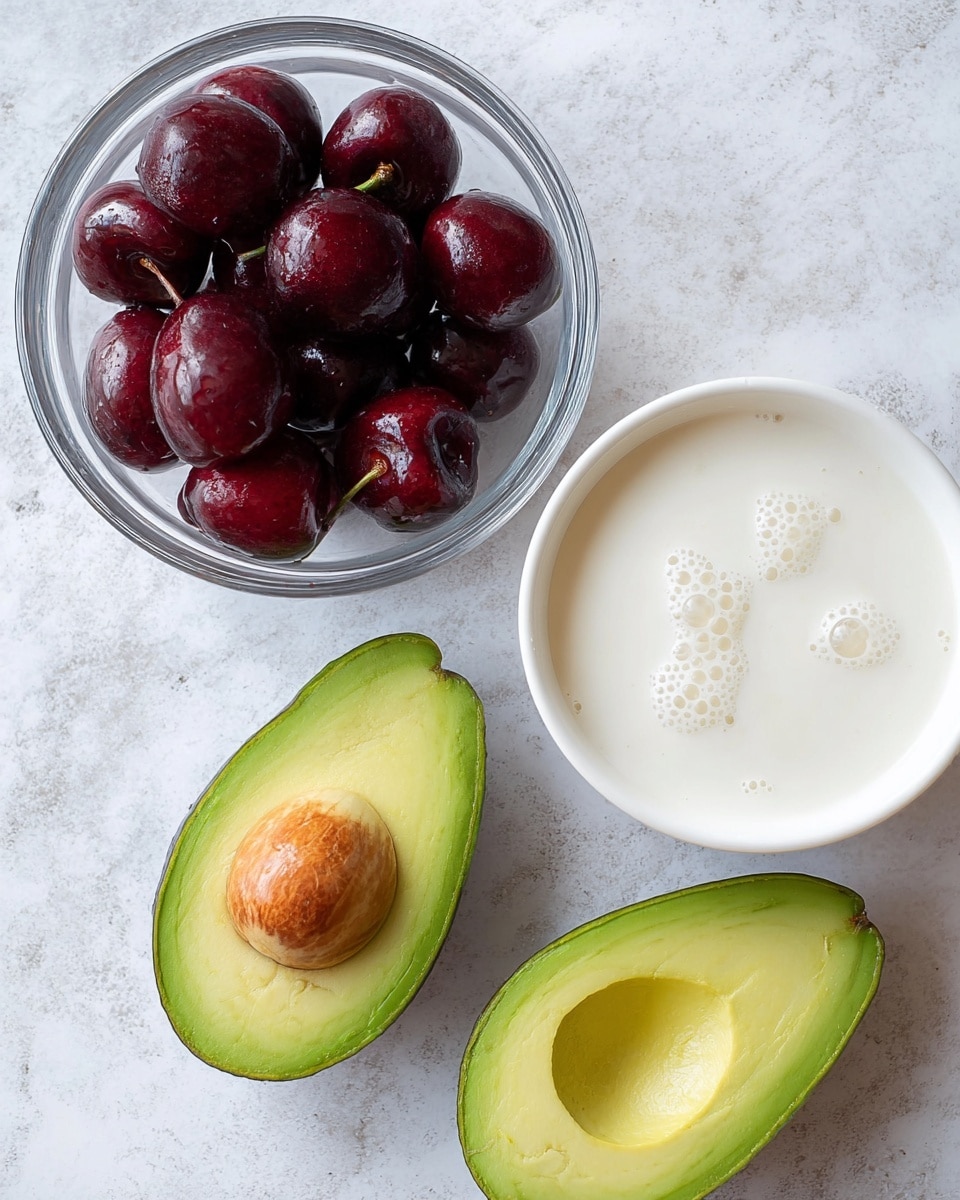 The image shows three main items on a white marbled surface: two avocado halves with bright green flesh and one with a brown seed in the center, a clear glass bowl filled with shiny dark red cherries, and a white bowl filled with a smooth, creamy white liquid that has small bubbles on the surface. All items are arranged loosely in a triangular shape. photo taken with an iphone --ar 4:5 --v 7