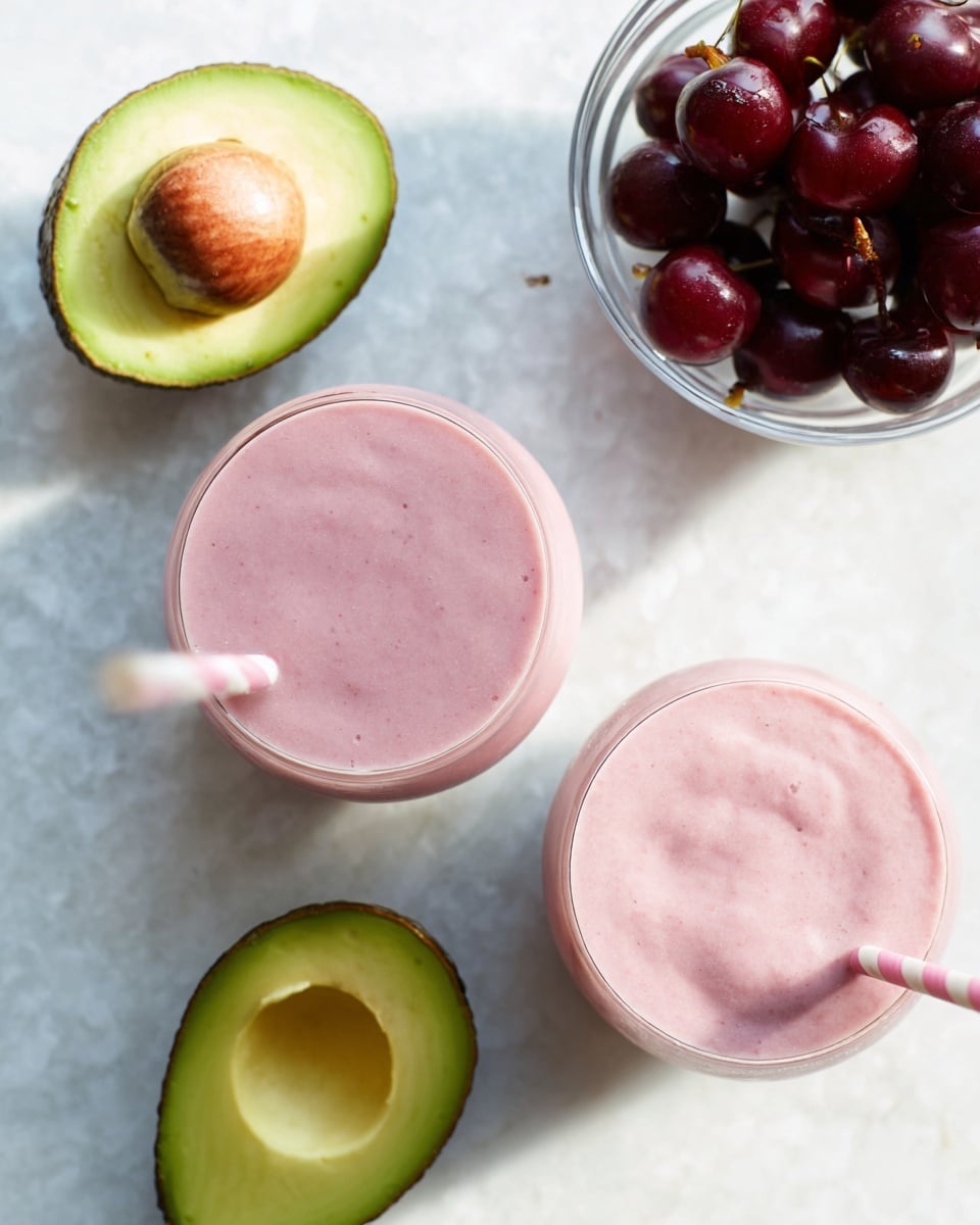 The image shows two round glasses filled with smooth, light pink smoothies, each with a white and pink striped straw. The glasses are placed on a white marbled surface. In the upper left corner, there is a half avocado displaying its green flesh and smooth texture. Below the glasses, another half avocado is seen with its brown seed intact and creamy green flesh. On the upper right side, a clear glass bowl contains dark red cherries with a shiny, fresh look. The overall scene uses soft natural light and soft shadows to highlight the colors and textures. Photo taken with an iphone --ar 4:5 --v 7
