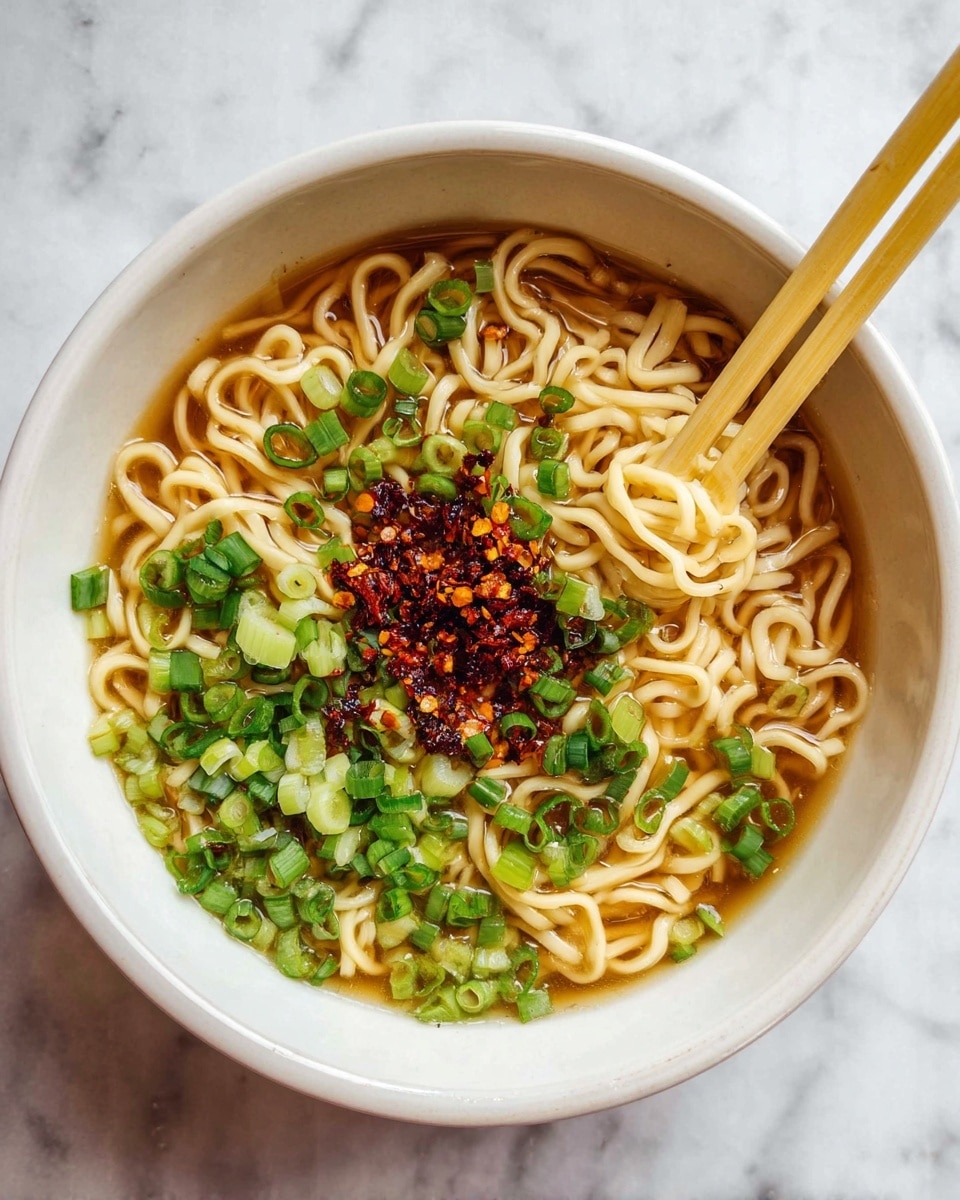 A white bowl filled with light brown noodles in broth, topped with bright green chopped spring onions all around the edges, and a small pile of chili flakes mixed with oil in the center. Light yellow chopsticks rest on the top edge of the bowl. The bowl sits on a white marbled surface. photo taken with an iphone --ar 4:5 --v 7