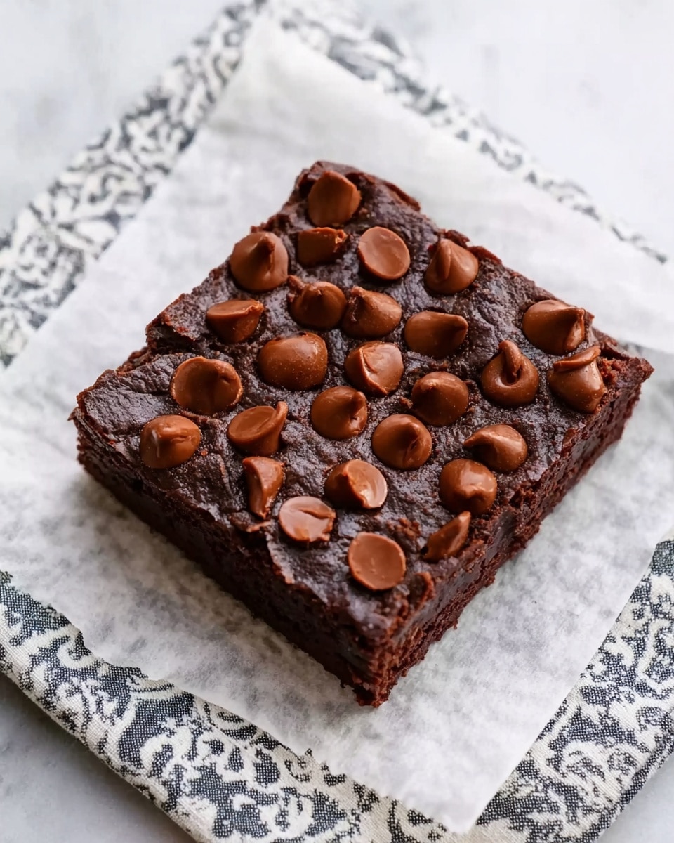 A single thick, square dark brown brownie with a rich, moist texture is placed on a piece of white parchment paper. The top of the brownie is covered evenly with large, smooth, and shiny milk chocolate chips that add a slightly rounded texture. The brownie is centered on a white marbled surface with a patterned cloth partially visible underneath the parchment paper. photo taken with an iphone --ar 4:5 --v 7