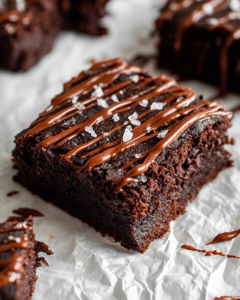A piece of dark brown chocolate brownie with a moist and dense texture, topped with smooth, shiny chocolate drizzle in spaced lines across the top, sprinkled with small crystals of sea salt. The brownie sits on crumpled white parchment paper, with some chocolate smudges near its edges, and part of another similar brownie is visible in the background on a white marbled surface. photo taken with an iphone --ar 4:5 --v 7