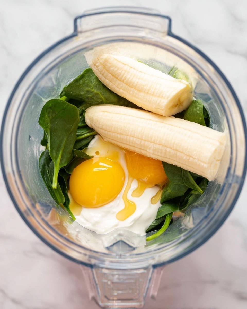 Inside a clear blender jar on a white marbled surface, there are two peeled bananas placed next to each other on top. Below them, two raw egg yolks with bright yellow color rest on a bed of fresh, dark green spinach leaves. There is also a dollop of creamy white yogurt mixed in towards the side, with a drizzle of golden honey visible on the yogurt's surface. The textures inside the blender vary from smooth and soft yogurt to the firm and fresh spinach leaves, creating a layered mix of colors from yellow to green to white. photo taken with an iphone --ar 4:5 --v 7