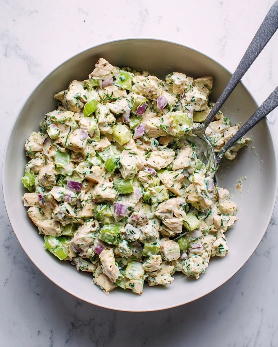 A large white bowl filled with a chunky salad made of small pieces of light beige chicken, pale green celery, and bits of purple onion all mixed with a creamy white dressing speckled with green herbs. Two gray-handled spoons rest inside the bowl on a white marbled surface. Photo taken with an iphone --ar 4:5 --v 7