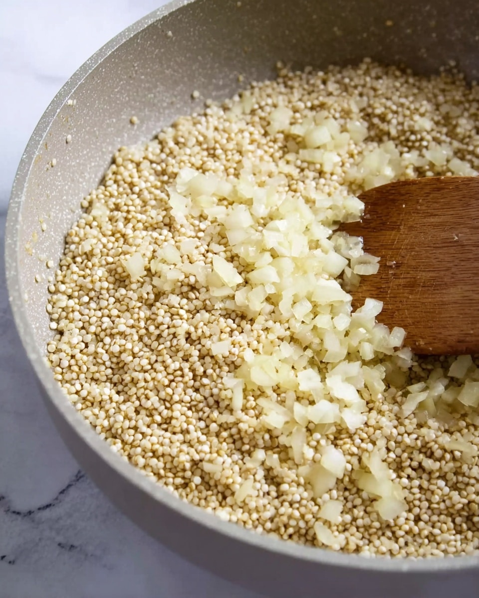 The image shows a close-up of a pan with two main layers. The bottom layer is a light beige grain, possibly quinoa, with a round, bead-like texture spread evenly across the pan. The top layer consists of small, finely chopped pale yellow pieces, likely onions, scattered unevenly over the grains, mainly on the right side. The pan itself is light gray and the cooking is being stirred by a wooden spatula visible from the bottom center of the image. The background is a white marbled texture. photo taken with an iphone --ar 4:5 --v 7