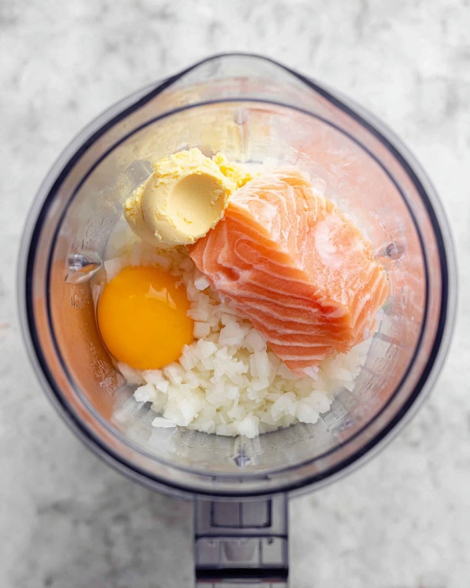 Inside a clear blender pitcher, there are several ingredients layered together. On the bottom, a thick layer of white chopped onions fills most of the space. Above the onions, there is a smooth, pale yellow dollop of minced garlic. Next to the garlic, a raw egg yolk with its bright orange color is visible. A raw piece of pink salmon rests on top, showing fine white lines of fat. The mixture is placed on a white marbled surface. Photo taken with an iphone --ar 4:5 --v 7