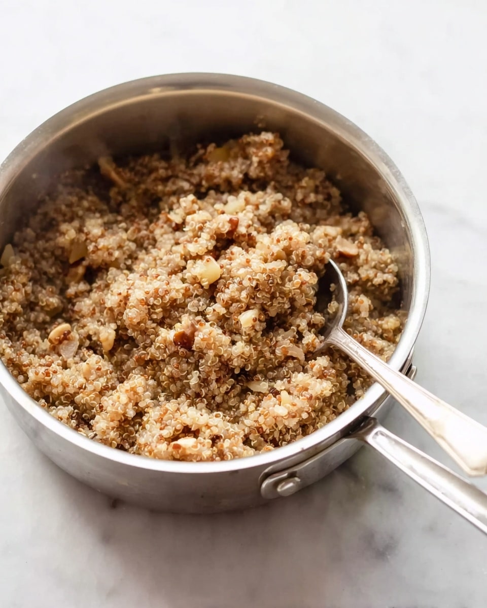 A close-up view of a silver metal saucepan filled with cooked quinoa mixed with small pieces of light brown and beige vegetables or nuts, creating a textured, slightly chunky mixture. The quinoa appears soft and fluffy with a warm, light brown color mixed with specks of beige. A silver spoon is resting inside the pan, partially submerged in the quinoa. The pan is placed on a white marbled surface. Steam rises gently from the dish, suggesting it is hot and fresh. photo taken with an iphone --ar 4:5 --v 7