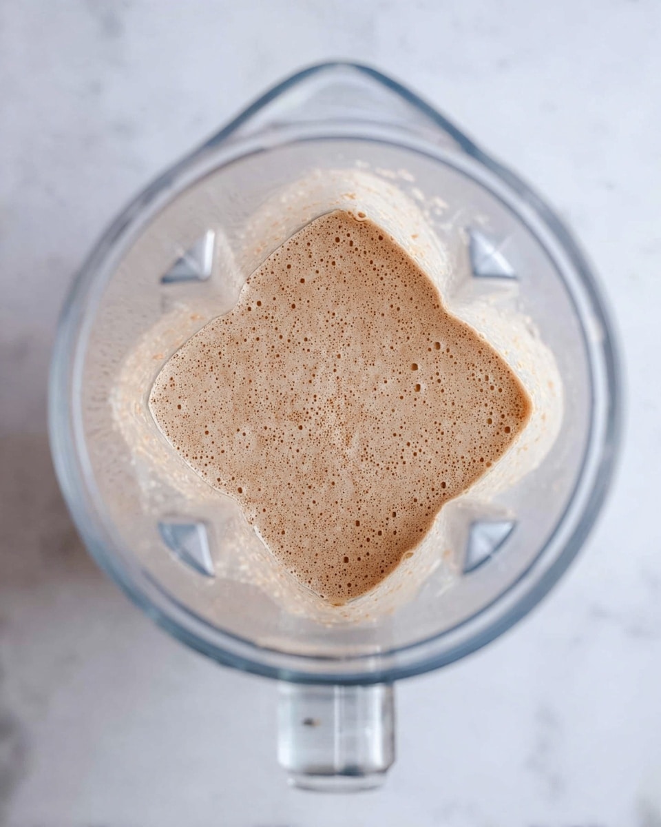 The image shows a top view of a clear blender container sitting on a white marbled surface. Inside the blender, there is a light brown mixture with small bubbles and a slightly frothy texture, filling only the bottom layer of the container. The sides of the blender are clean, and the container’s handle is visible at the bottom of the image. Photo taken with an iphone --ar 4:5 --v 7