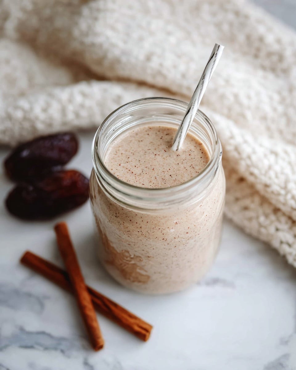 A clear glass jar filled with a light brown creamy drink speckled with tiny dark particles. Inside the jar, a white straw with thin black lines leans slightly to the right. The jar stands on a white marbled surface with two cinnamon sticks placed nearby on the lower right side. Behind the jar, there is a soft, off-white knitted fabric that adds texture to the background. In the upper left corner, two dark brown dates are partially visible, resting on the same surface. The photo taken with an iphone --ar 4:5 --v 7