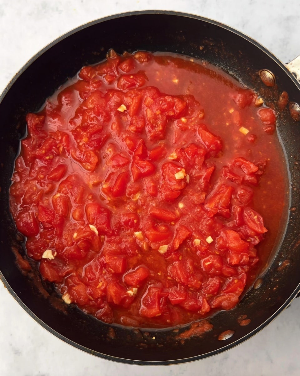 The image shows a black pan filled with a chunky red tomato sauce. The sauce has visible pieces of diced tomatoes with a soft texture, floating in a thin, bright red tomato liquid. Small bits of white minced garlic or onion are mixed evenly throughout the sauce, adding texture. The sauce looks thick in some parts and watery in others, covering the bottom of the pan in a single layer. The pan sits on a white marbled surface. photo taken with an iphone --ar 4:5 --v 7