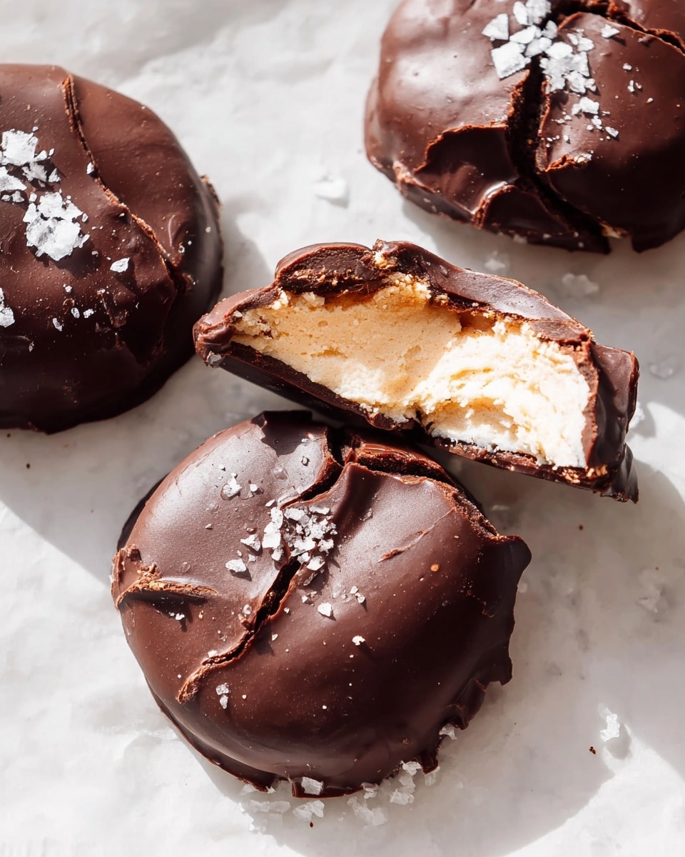 The image shows three round chocolate-covered treats on a white marbled surface, with one in the center partly broken open. Each treat has two visible layers: a smooth dark brown chocolate outer layer that looks thick and slightly crackled on top, and a creamy pale beige filling inside that appears soft and fluffy. The top chocolate layer on the broken treat is cracked in pieces, revealing the light, airy texture of the filling. Scattered sea salt flakes rest on the chocolate surface, adding a rough white texture contrast. The lighting accentuates the shine on the chocolate and the soft texture of the filling. photo taken with an iphone --ar 4:5 --v 7