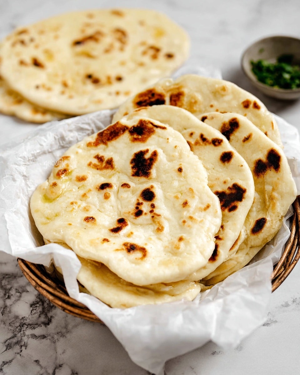 A basket lined with white paper holds a stack of five round flatbreads with light golden color and dark brown char marks scattered unevenly across their slightly puffed, soft texture. Behind the basket, there is one flatbread lying flat on a white marbled surface, showing similar browning. In the background, a small bowl filled with green herbs is visible, adding a touch of color. The overall setting is clean and bright with a white marbled texture under and around the basket. photo taken with an iphone --ar 4:5 --v 7