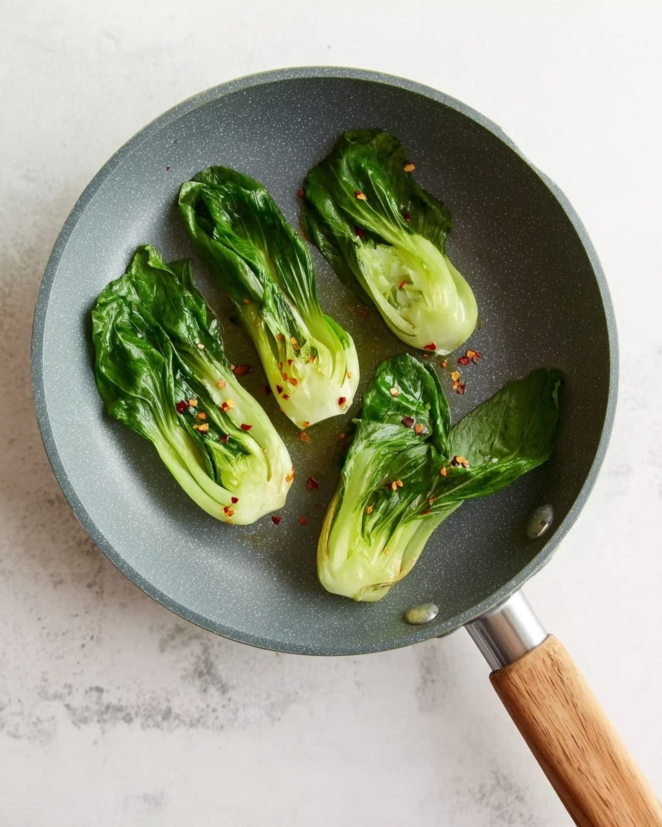 A gray frying pan with a wooden handle contains four baby bok choy pieces cooked lightly. The bok choy is arranged with two pieces on the left side, one larger piece near the top center, and one smaller piece near the center right. The bok choy leaves are a deep green color with the stalks being light green to pale white. Small red chili flakes are sprinkled over the bok choy, adding a touch of color contrast. The pan sits on a white marbled surface. photo taken with an iphone --ar 4:5 --v 7