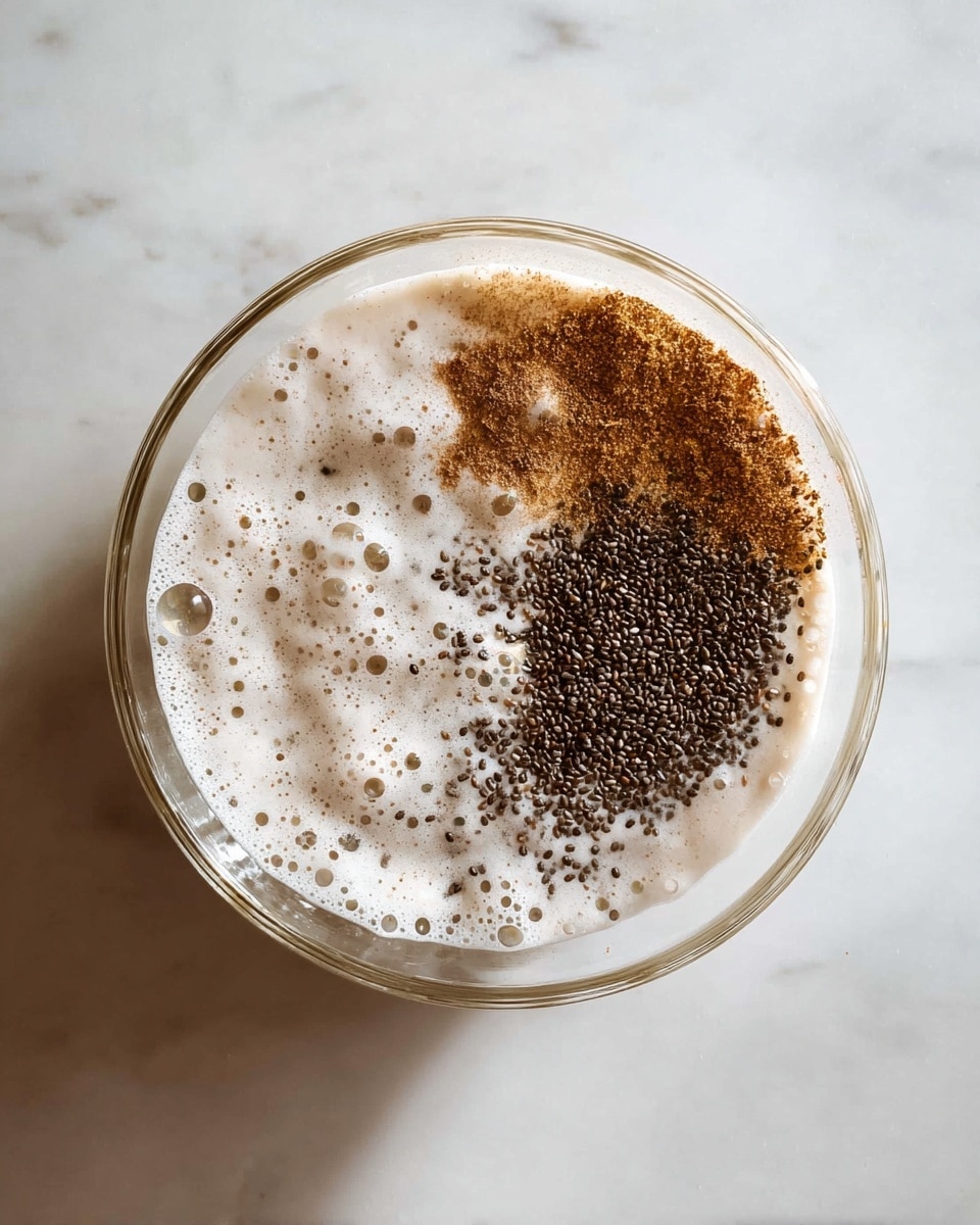 A clear glass bowl sits on a white marbled surface, filled with a mix of creamy white liquid and dry ingredients separated on top. The liquid layer is frothy with bubbles, while on its surface there is a dark brown pile of tiny chia seeds on the right side, scattered brown powder in the center and left, and some small black specks spread throughout. The scene is bright with natural light showing the texture of the ingredients clearly photo taken with an iphone --ar 4:5 --v 7