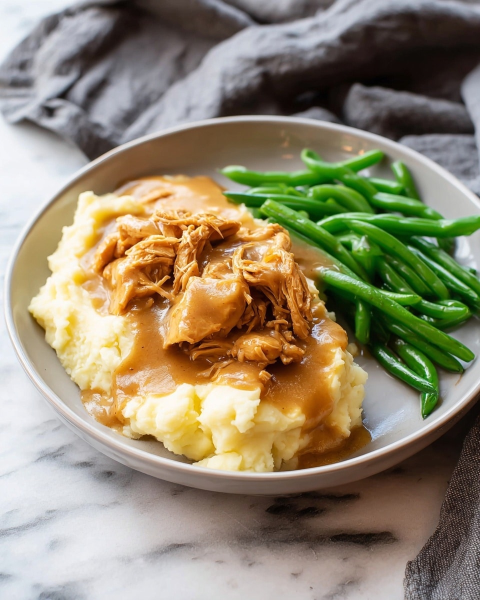 The image shows a white plate with three main layers of food. On the left side, there is a base layer of creamy mashed potatoes in a smooth, soft texture with a pale yellow color. On top of the mashed potatoes, a layer of shredded, brownish-orange chicken pieces covered in thick, light brown gravy spreads over the potatoes and slightly onto the plate. On the right side of the plate, bright green, fresh green beans with a smooth texture are placed next to the mashed potatoes and chicken. The plate is on a white marbled surface, and a soft, grey cloth lies nearby. Photo taken with an iphone --ar 4:5 --v 7