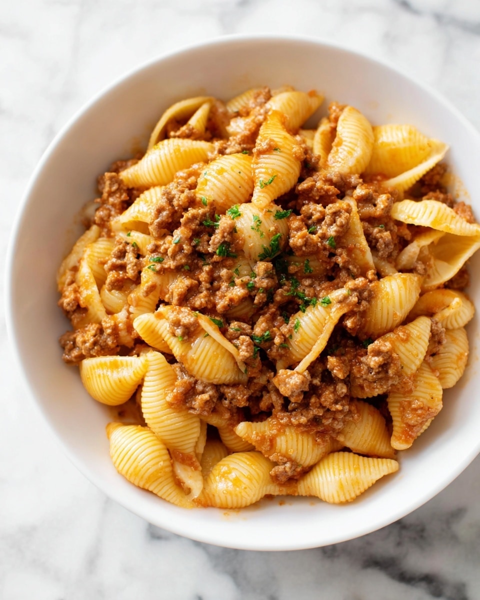 A white bowl filled with three layers of food: the bottom and middle layer are made of small shell pasta that are pale yellow with ridges, coated lightly in a sauce. The top layer is a mix of cooked ground meat pieces in a reddish-brown sauce with small bits of onion and herbs sprinkled on top, adding small green flecks. The pasta and meat are well mixed, and the surface looks creamy and warm. The bowl sits on a white marbled surface. Photo taken with an iphone --ar 4:5 --v 7