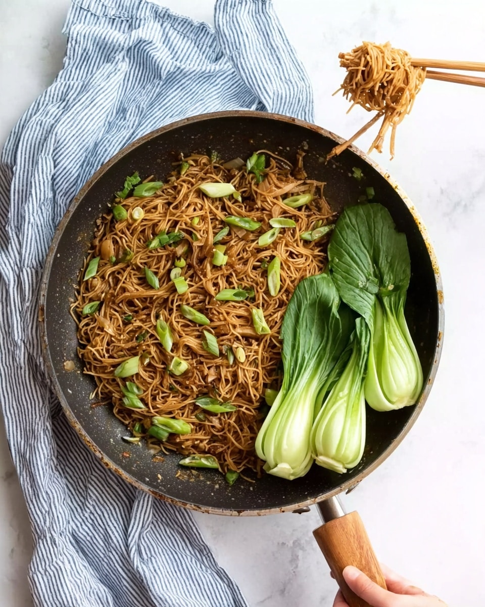 A pan filled with thin brown noodles mixed with small green onion pieces and some light brown bits, with two bunches of bright green bok choy placed on the right side of the pan. A woman’s hand holding two wooden chopsticks is lifting a small bundle of noodles above the pan. The pan has a dark interior and a wooden handle and is placed on a white marbled surface with a striped blue and white cloth napkin beside it. photo taken with an iphone --ar 4:5 --v 7
