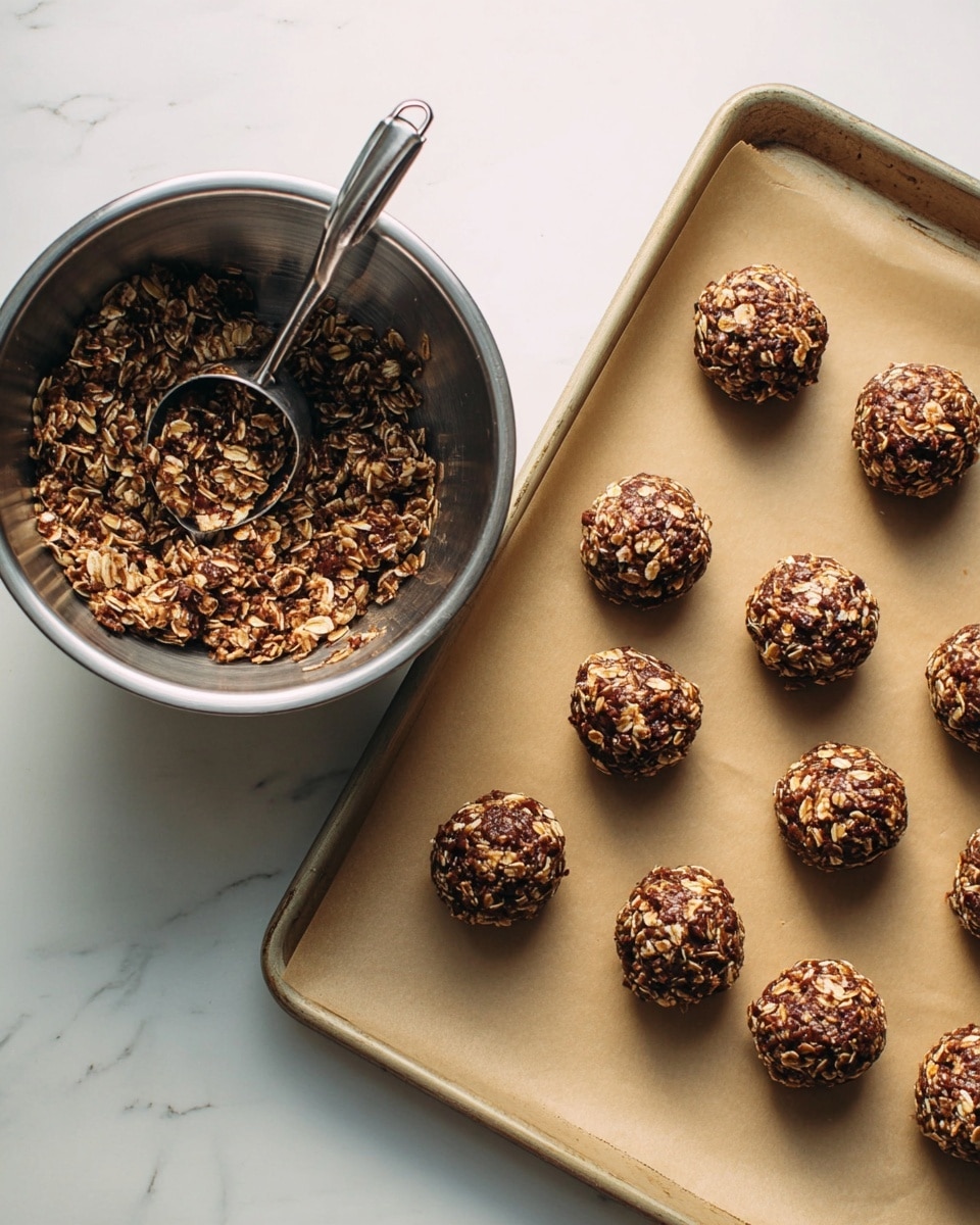 This image shows a baking tray lined with light brown parchment paper holding twelve rounded cookie dough balls, each made from a mixture of oats and chocolate, giving them a textured, chunky look with dark brown and light tan colors. To the left, there is a shiny metal mixing bowl filled with the same oatmeal and chocolate dough mixture, with a metal scoop resting inside. The whole setup is placed on a white marbled surface. photo taken with an iphone --ar 4:5 --v 7