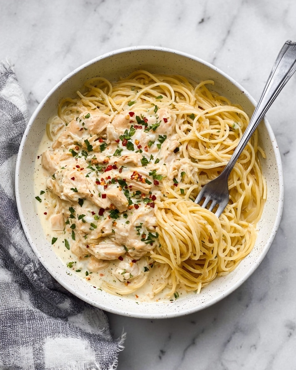 A white speckled bowl filled with thin, light yellow spaghetti noodles forming the bottom layer, topped with a creamy, thick off-white sauce that covers pieces of light beige cooked chicken. The sauce is garnished with small bits of green herbs and tiny red chili flakes scattered on top. A silver fork pierces into the sauce and noodles, gathering a small bundle of the spaghetti. The bowl rests on a white marbled surface with a gray and white checkered cloth partially visible on the side. Photo taken with an iphone --ar 4:5 --v 7