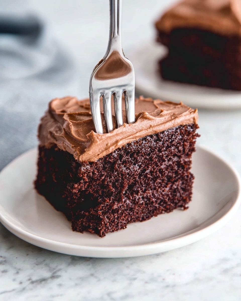 A piece of rich dark brown chocolate cake with a thick layer of smooth, creamy light brown chocolate frosting on top sits on a white plate. A shiny silver fork is pressing gently into the frosting on the top center of the cake. The cake looks soft and moist with a textured crumb. A blurred second piece of cake is visible in the background on another white plate. The surface underneath is a white marbled texture. photo taken with an iphone --ar 4:5 --v 7