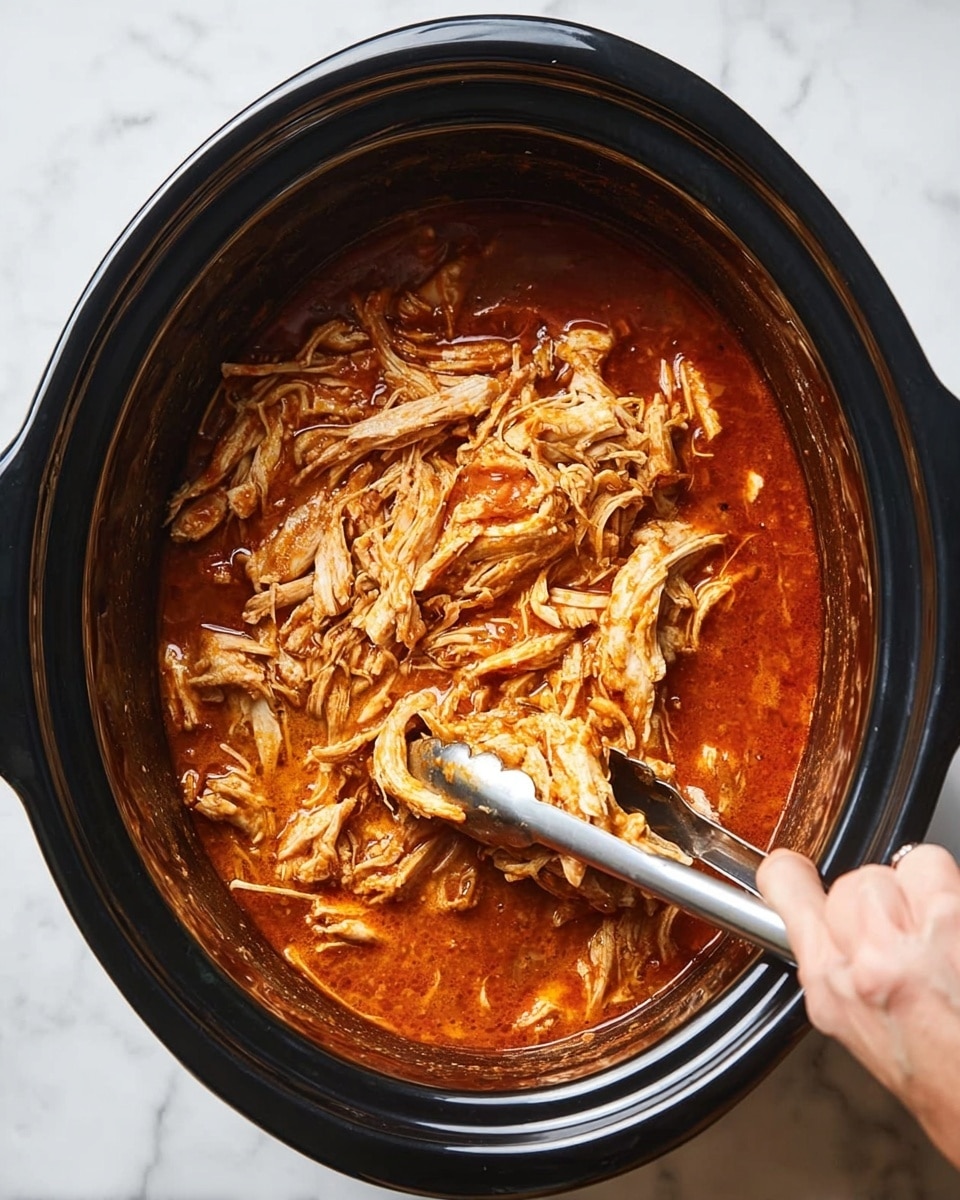 A black slow cooker is filled with shredded chicken in a thick, reddish-orange sauce. The chicken is pulled into small and medium pieces with visible white and light brown strands mixing with the rich sauce that has a glossy texture. A pair of silver tongs held by a woman's hand is shown lifting some of the shredded chicken. The cooker sits on a white marbled surface. photo taken with an iphone --ar 4:5 --v 7