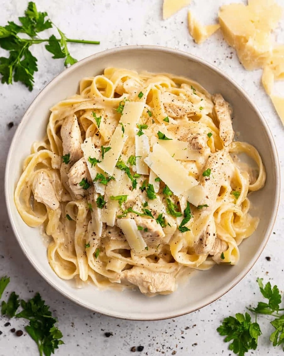 A white bowl filled with creamy fettuccine pasta in a light beige sauce, topped with pieces of tender white chicken. The pasta is mixed with thin, flat noodles that have a smooth texture and are coated evenly with sauce. On top, there are scattered thin slices of pale yellow cheese and sprinkled chopped green parsley, adding fresh color. Small black pepper spots are visible on the chicken pieces. The bowl is placed on a white marbled surface with some parsley leaves and cheese shards around it. photo taken with an iphone --ar 4:5 --v 7