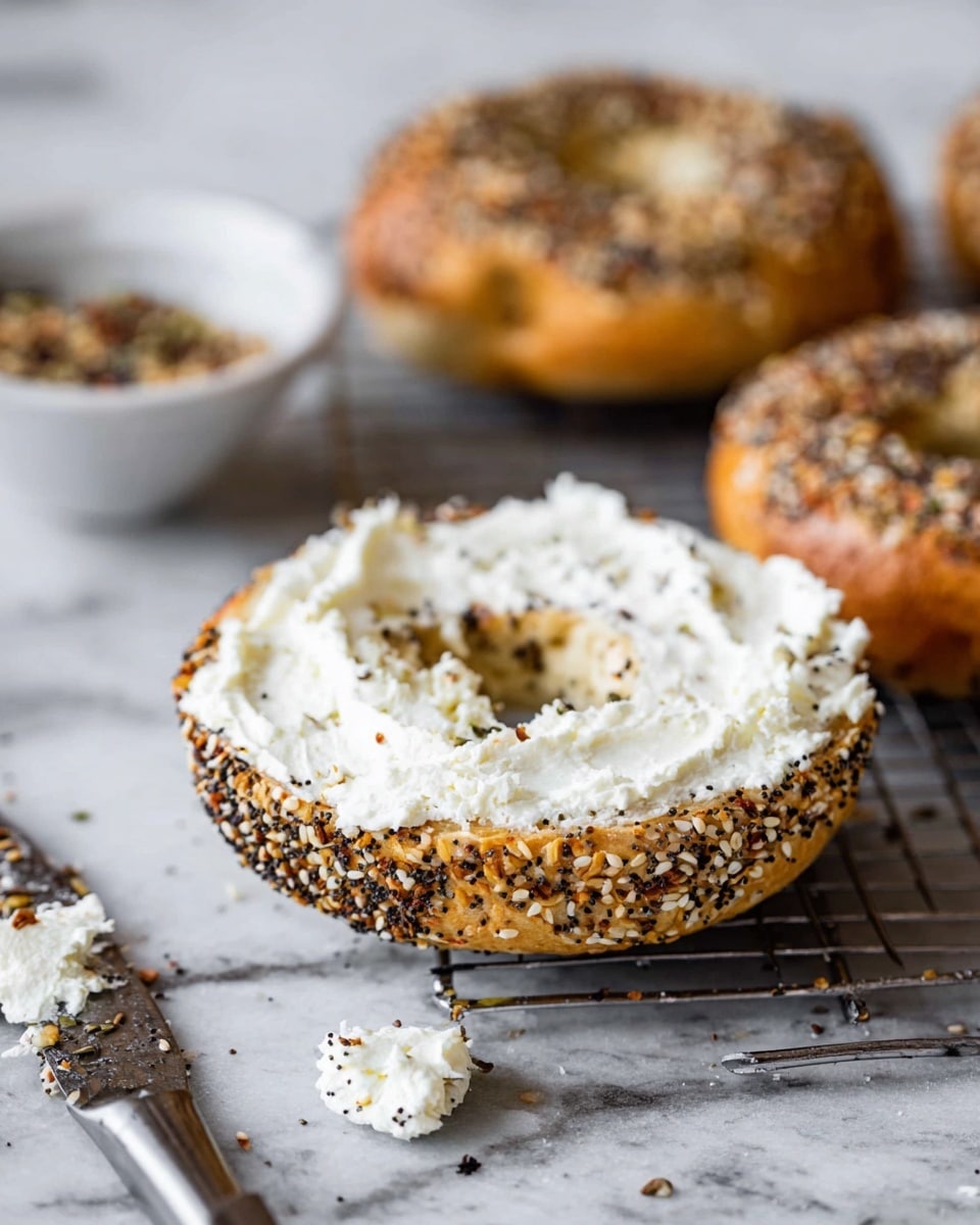 The image shows a close-up of an everything bagel sliced in half, with the bottom half spread thickly with creamy white cream cheese. The bagel is golden brown with a crispy texture and is covered with seeds like sesame, poppy, and onion, creating a speckled pattern. The top half of the bagel is in the background, slightly out of focus, also showing the seeded crust. The bagel rests on a silver cooling rack over a white marbled surface. Nearby, there is a white bowl with a mixture of seeds and spices, and a knife with some cream cheese on the blade lies on the surface. Photo taken with an iphone --ar 4:5 --v 7