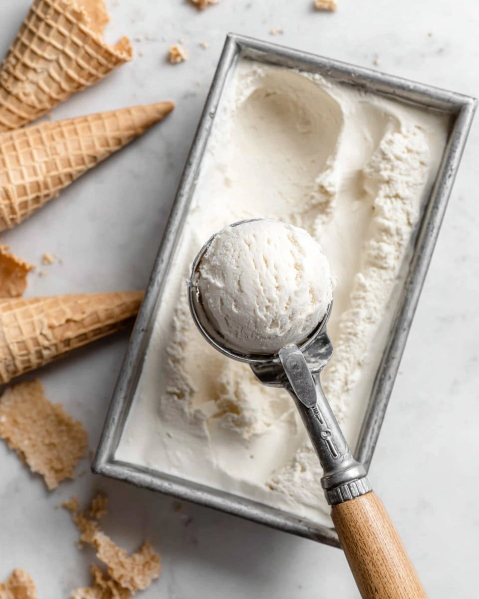 A close-up view of a single scoop of creamy white ice cream held in a silver ice cream scooper with a wooden handle, positioned above a metal tray filled with smooth, spread ice cream of the same white color and creamy texture. To the left of the tray, there are two empty white waffle cones stacked one on top of the other, and scattered waffle pieces around them. The whole scene is set on a white marbled surface. photo taken with an iphone --ar 4:5 --v 7