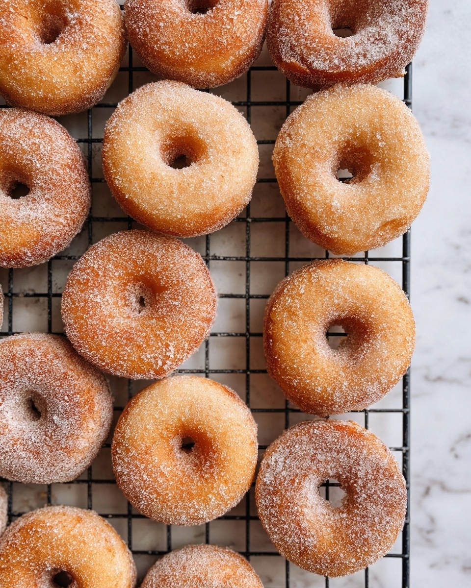 The image shows many small, round doughnuts arranged closely together on a black wire cooling rack. Each doughnut has a golden-brown color with a dusting of fine white sugar on the surface, giving a slightly textured look. They all have a small hole in the center and look soft and fluffy. The cooling rack is placed on a white marbled surface which adds a clean and bright background to the warm tones of the doughnuts. Photo taken with an iphone --ar 4:5 --v 7
