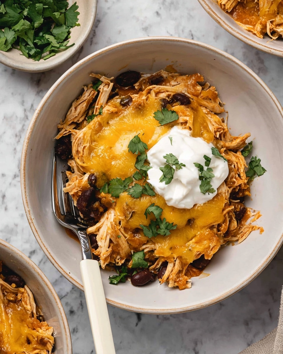 A white bowl holds a layered dish with shredded light brown chicken mixed with black beans as the base. Over the top, several layers of melted yellow cheese partially cover the chicken, giving it a slightly shiny look. At the center, there is a dollop of white sour cream, garnished with small, bright green chopped cilantro leaves scattered around. A fork with a white handle rests on the left side inside the bowl. The bowl sits on a white marbled surface with another similar white bowl partially visible. Photo taken with an iphone --ar 4:5 --v 7