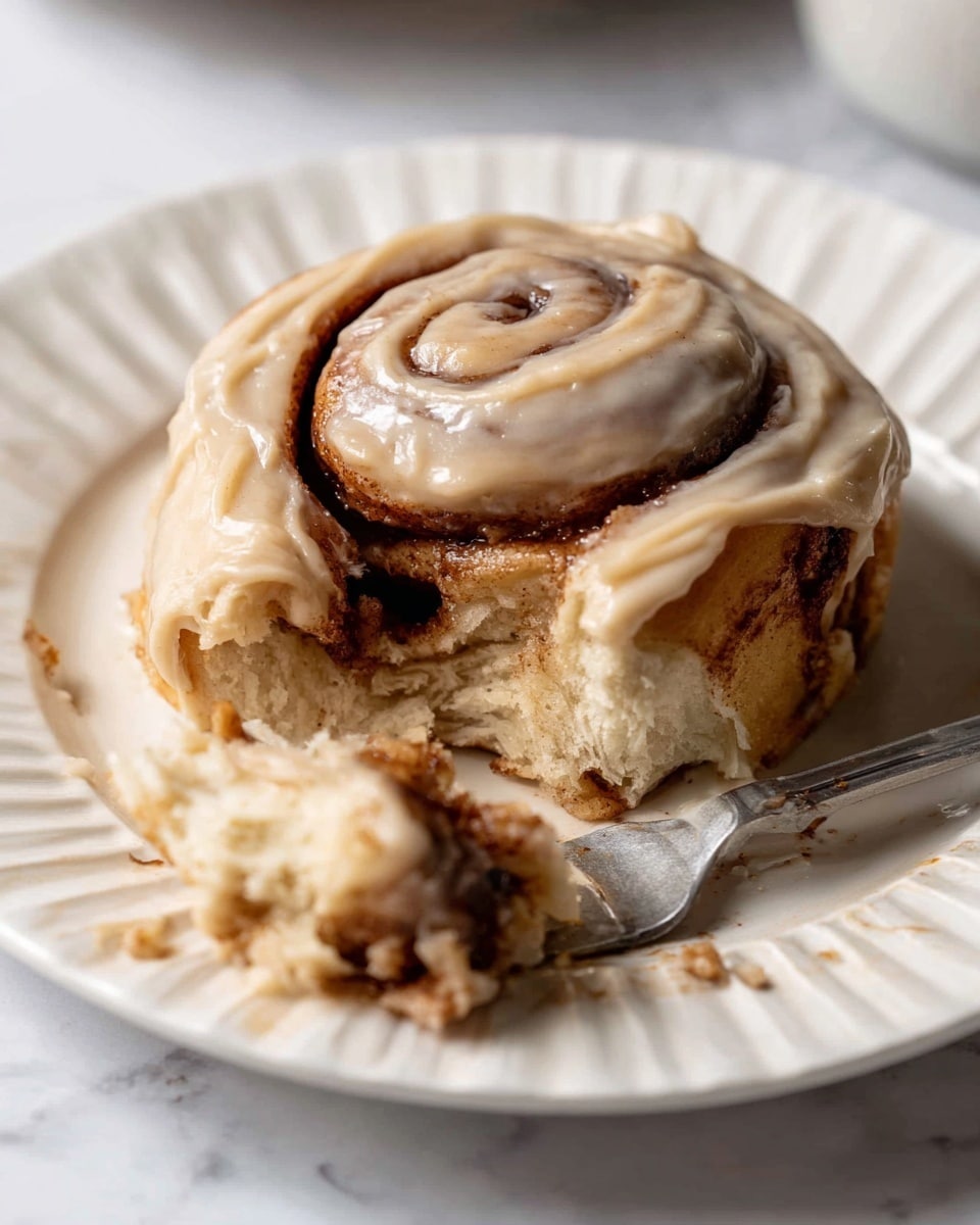 A single cinnamon roll sits on a white fluted plate placed on a white marbled surface. The cinnamon roll has three visible spiraled layers, with the inner layers showing a dark brown cinnamon filling and the outer dough a light golden color. A thick, creamy light tan frosting generously covers the top and slightly drapes over the sides of the roll. A small piece of the roll has been taken off and is resting on a fork in front of the main roll, revealing the soft, fluffy texture of the dough and the cinnamon filling inside. The scene is well-lit, showing a smooth and moist texture on the frosting and a tender look to the roll. Photo taken with an iphone --ar 4:5 --v 7