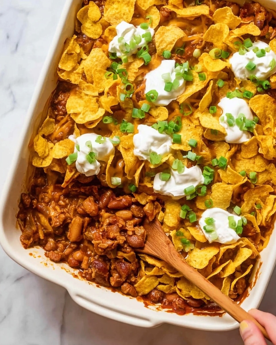 The dish is in a white rectangular baking dish on a white marbled surface. It has three main layers: the bottom layer is a mix of cooked beans and small pieces of chicken in a thick reddish-brown sauce with visible spices. The middle layer is crispy yellow corn chips scattered evenly, covering most of the beans and chicken. The top layer has dollops of white sour cream and chopped green onions sprinkled over the chips. A wooden spoon held by a woman's hand is scooping some of the beans, sauce, and chips from one corner. Photo taken with an iphone --ar 4:5 --v 7