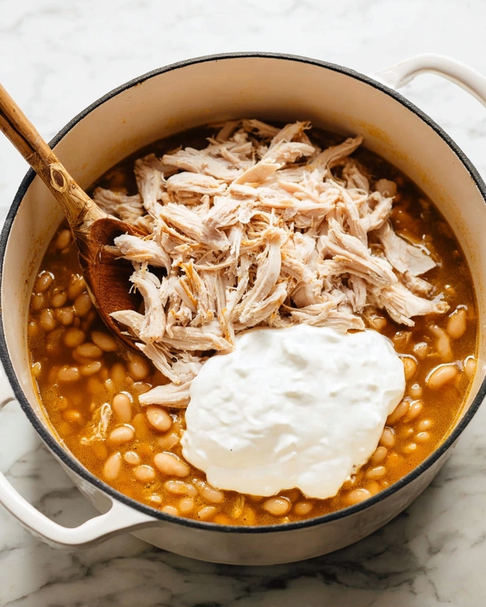 A white cooking pot filled with three main visible layers: the bottom layer is a golden-brown broth with small beans, the middle layer consists of shredded light tan meat pieces scattered over the beans, and the top layer is a dollop of thick, creamy white sour cream resting in the center. A wooden spoon with a light brown handle is placed inside the pot on the left side, partially submerged in the ingredients. The pot sits on a white marbled surface. Photo taken with an iphone --ar 4:5 --v 7