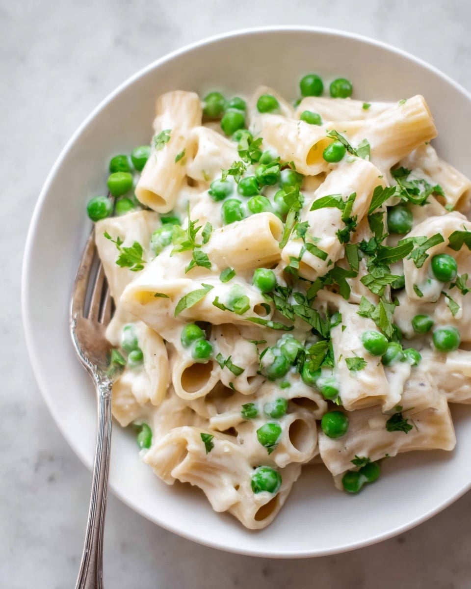 A white bowl holds one layer of short tube pasta coated in white creamy sauce, topped with bright green peas scattered evenly across the pasta, along with small pieces of fresh green herbs finely chopped and sprinkled on top. A silver fork rests inside the bowl, partially visible to the left. The bowl sits on a white marbled surface. photo taken with an iphone --ar 4:5 --v 7