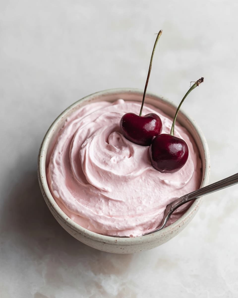 A small round bowl filled with one smooth, thick layer of pale pink whipped cream with soft swirls on the surface, two dark red cherries with long green stems placed on top for decoration, and a metal spoon inserted into the cream from the bottom right of the bowl, all set on a white marbled surface. photo taken with an iphone --ar 4:5 --v 7