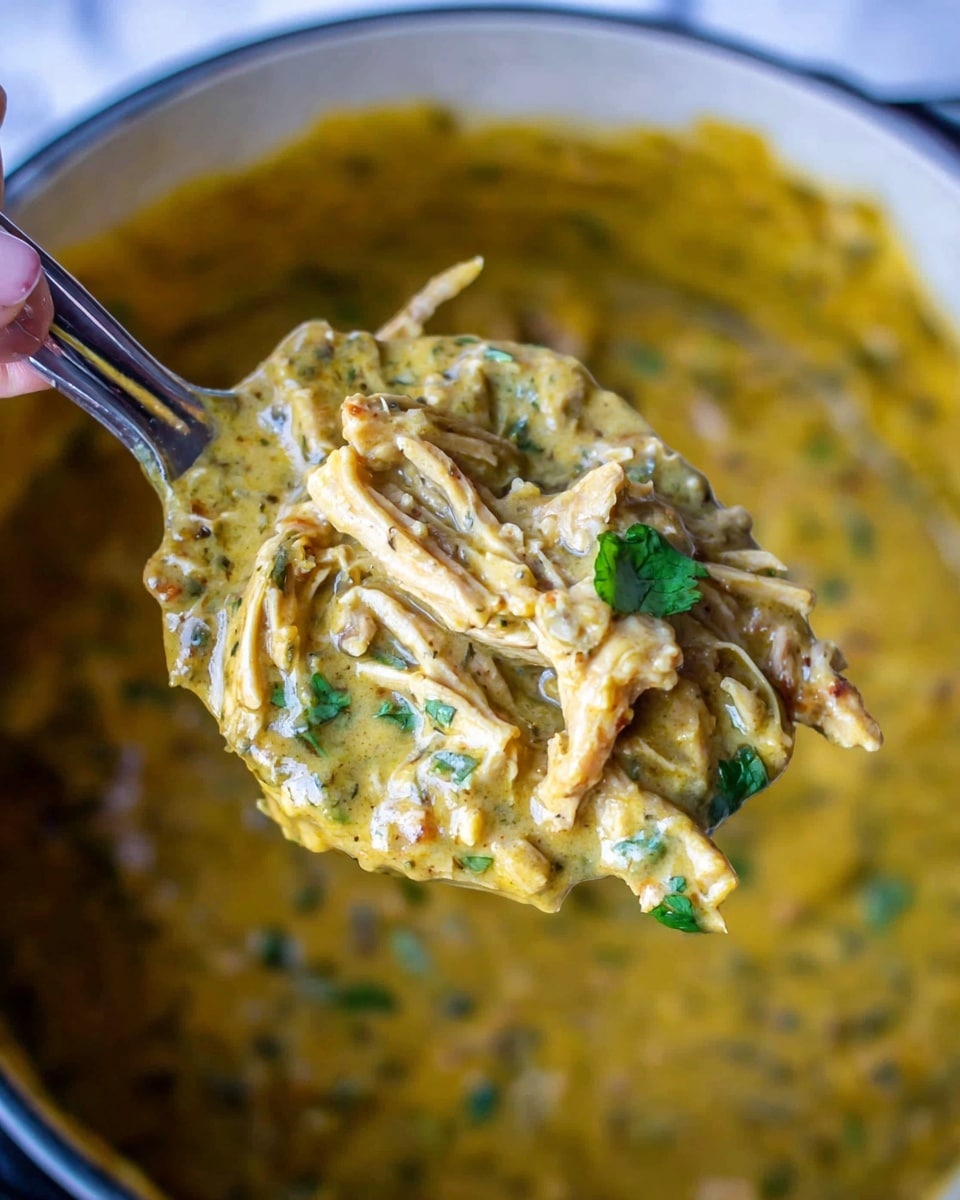 The image shows a close-up of a spoon scooping a thick creamy stew from a pot. The stew has several layers: base layer is a smooth yellow sauce with herbs and spices giving a speckled effect, mixed with shredded pieces of light brown meat and small green cilantro leaves scattered throughout. The texture looks rich and smooth with the shredded meat pieces visibly soft and tender within the sauce. The stew fills the spoon fully with the mixture held together, captured against a blurred background of the same stew inside a large pot. A woman's hand is holding the spoon from the top left corner, with part of the metal spoon handle visible. The surface beneath the pot is white marbled. photo taken with an iphone --ar 4:5 --v 7