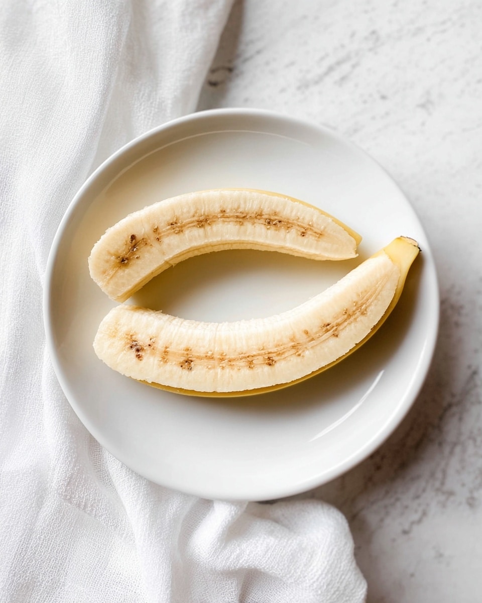 A white round plate holds two halves of a peeled banana, placed side by side with the inner, soft yellowish flesh and brown seeds showing clearly. The banana halves curve gently and rest flat on the plate, which sits on a white marbled surface with a softly wrinkled white cloth nearby. The image is bright and simple with soft natural lighting highlighting the banana's texture and the smooth plate. photo taken with an iphone --ar 4:5 --v 7