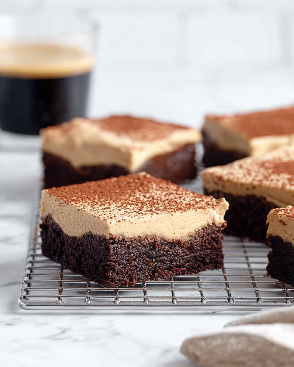The image shows several square chocolate brownies with a thick light brown creamy layer on top, sprinkled lightly with cocoa powder. Each brownie has two layers: the bottom layer is dark brown and dense with a slightly moist texture, and the top layer is smooth, light brown, and creamy, covering the brownies evenly. They are placed on a metal cooling rack set on a white marbled surface. In the background, there's a blurred glass cup of dark coffee. Photo taken with an iphone --ar 4:5 --v 7
