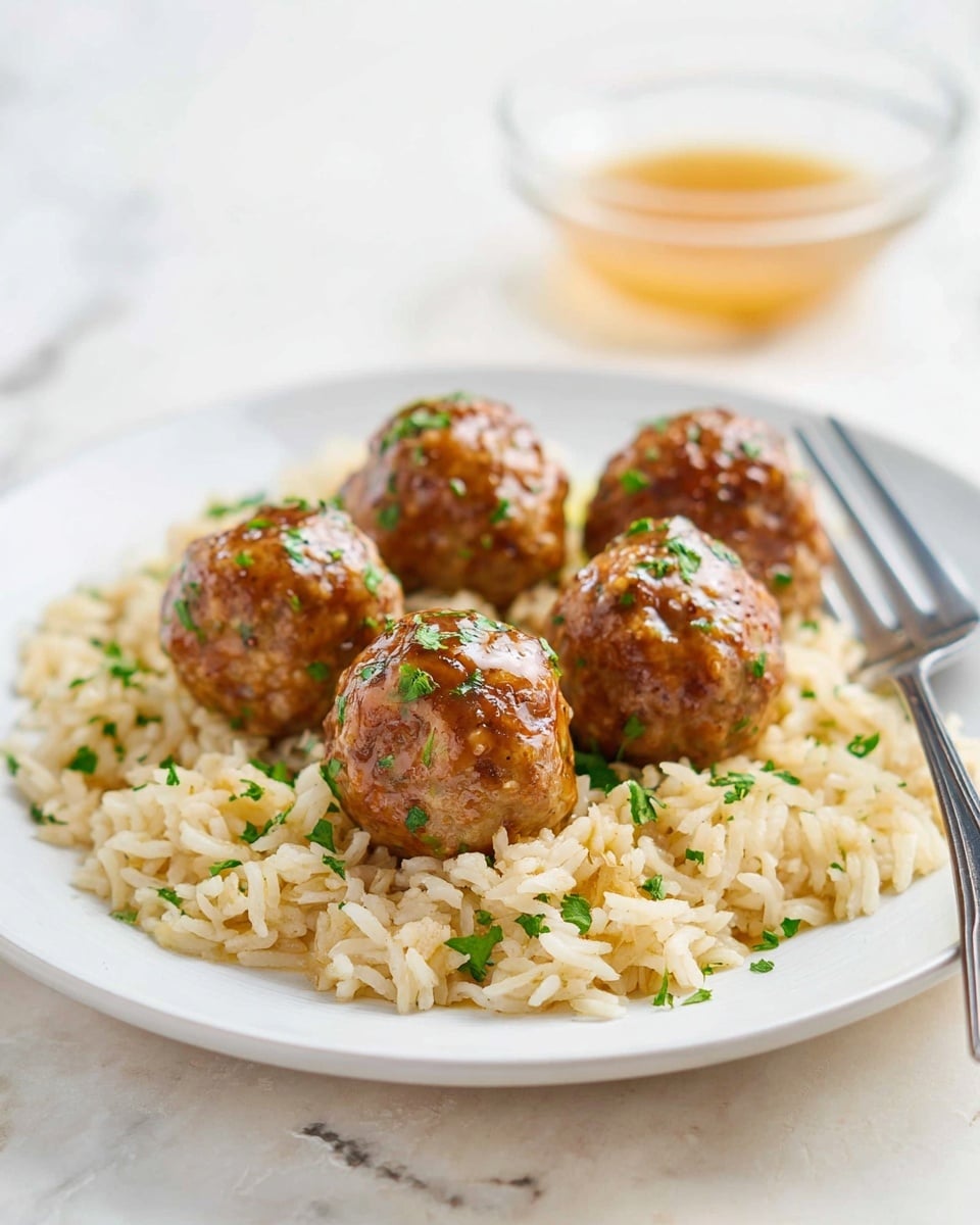 The image shows five shiny, brown meatballs with small green herb bits on them placed on a bed of light beige cooked rice on a white plate. The rice looks fluffy and also has some green herbs sprinkled on it. A metal fork rests on the edge of the plate behind the meatballs. In the blurred background, there is a small clear bowl with a light brown sauce. The whole setting is on a white marbled surface. photo taken with an iphone --ar 4:5 --v 7