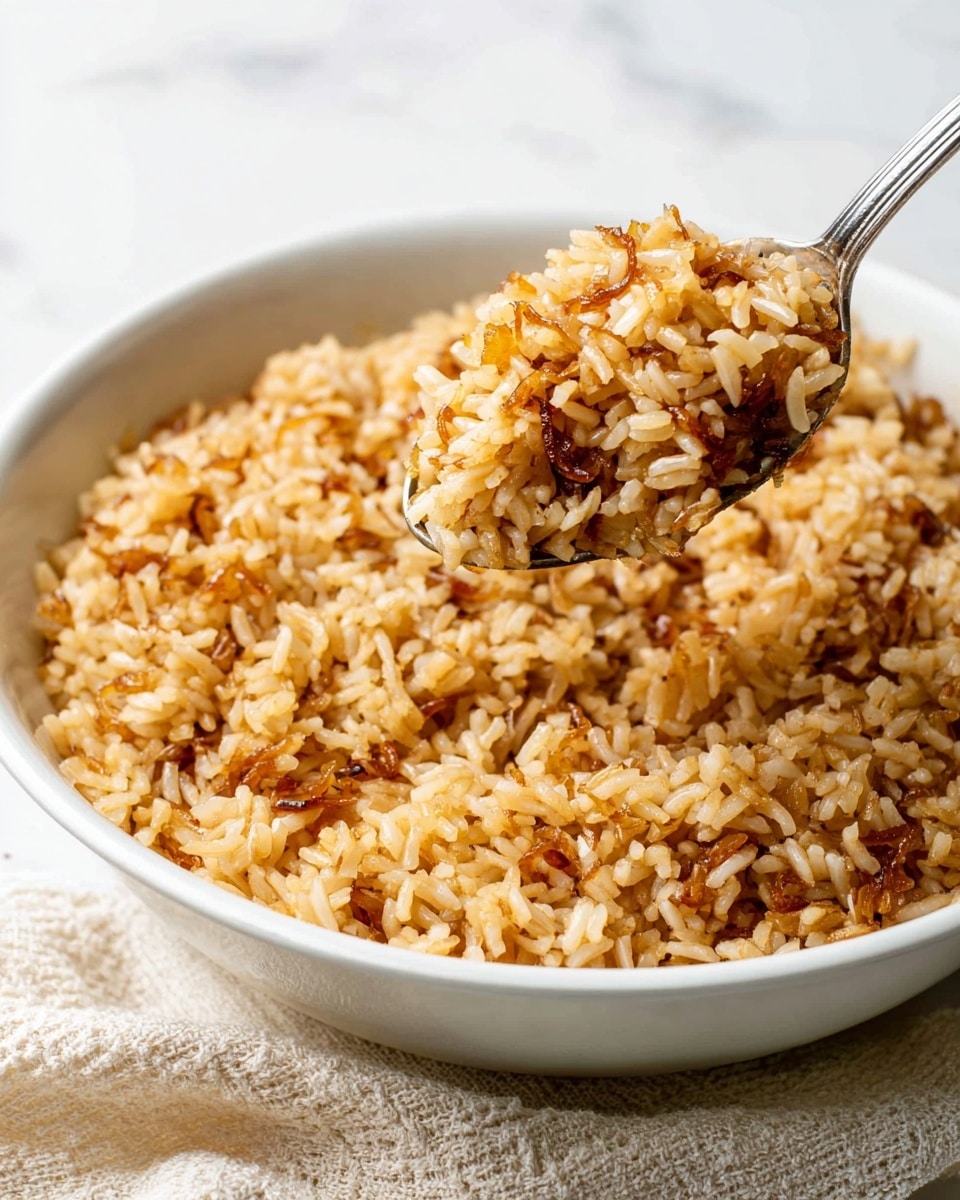 A large white bowl filled with cooked brown rice mixed with small pieces of caramelized onions. The rice looks fluffy but slightly sticky, with a golden brown color from cooking. A silver spoon holding a scoop of the rice hovers above the bowl, showing the texture and color of the grains and onion bits clearly. The background is a clean white marbled surface, and a beige textured cloth is partially visible at the front. photo taken with an iphone --ar 4:5 --v 7