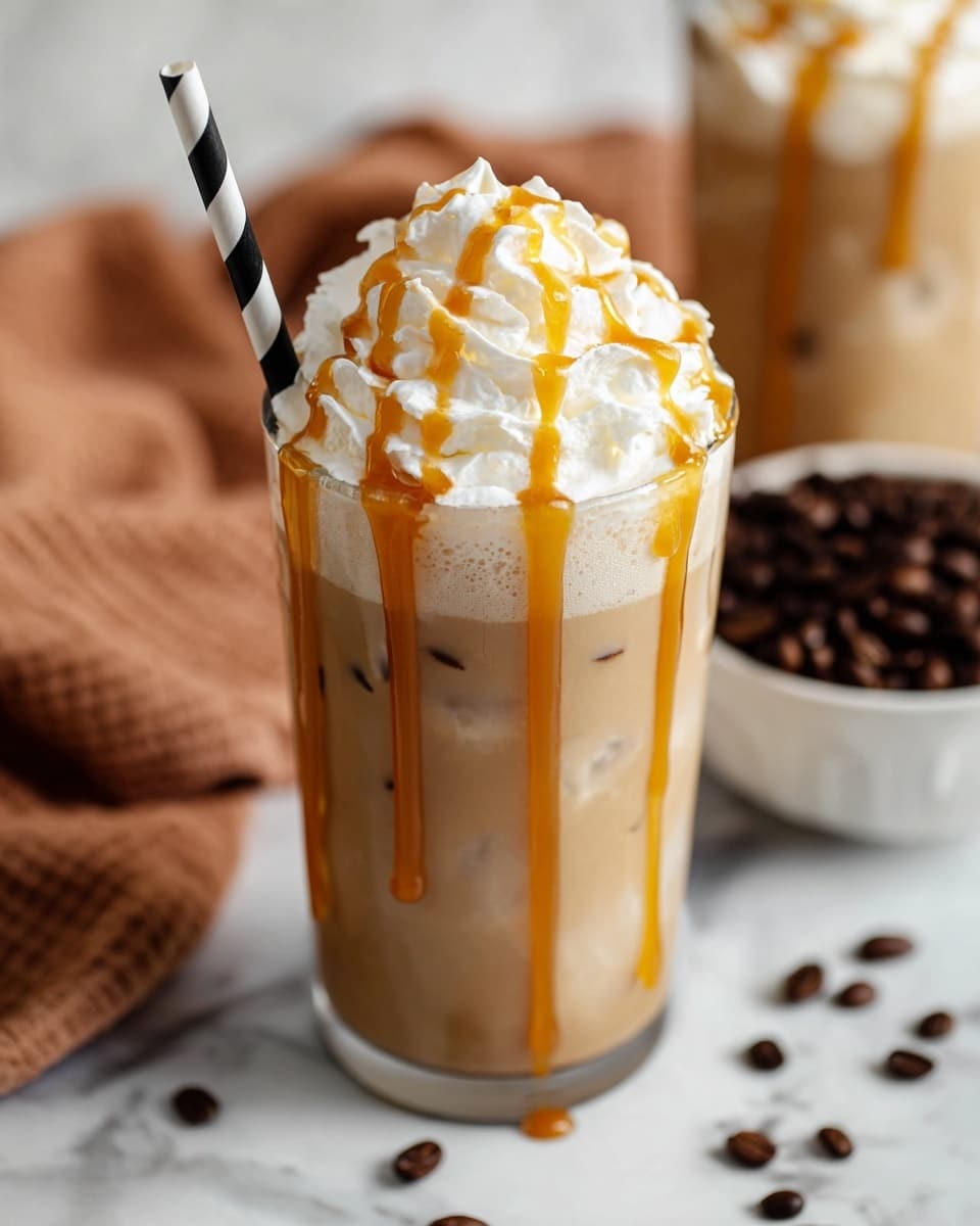 A clear glass filled with a light brown iced coffee drink topped with one thick layer of white whipped cream that has soft, swirled peaks. The whipped cream is drizzled generously with orange caramel sauce that also drips down the sides of the glass. A black and white striped paper straw is placed on the left side of the glass. In the background is a white bowl filled with dark roasted coffee beans, set against a white marbled surface with a blurred light brown cloth to the left. photo taken with an iphone --ar 4:5 --v 7