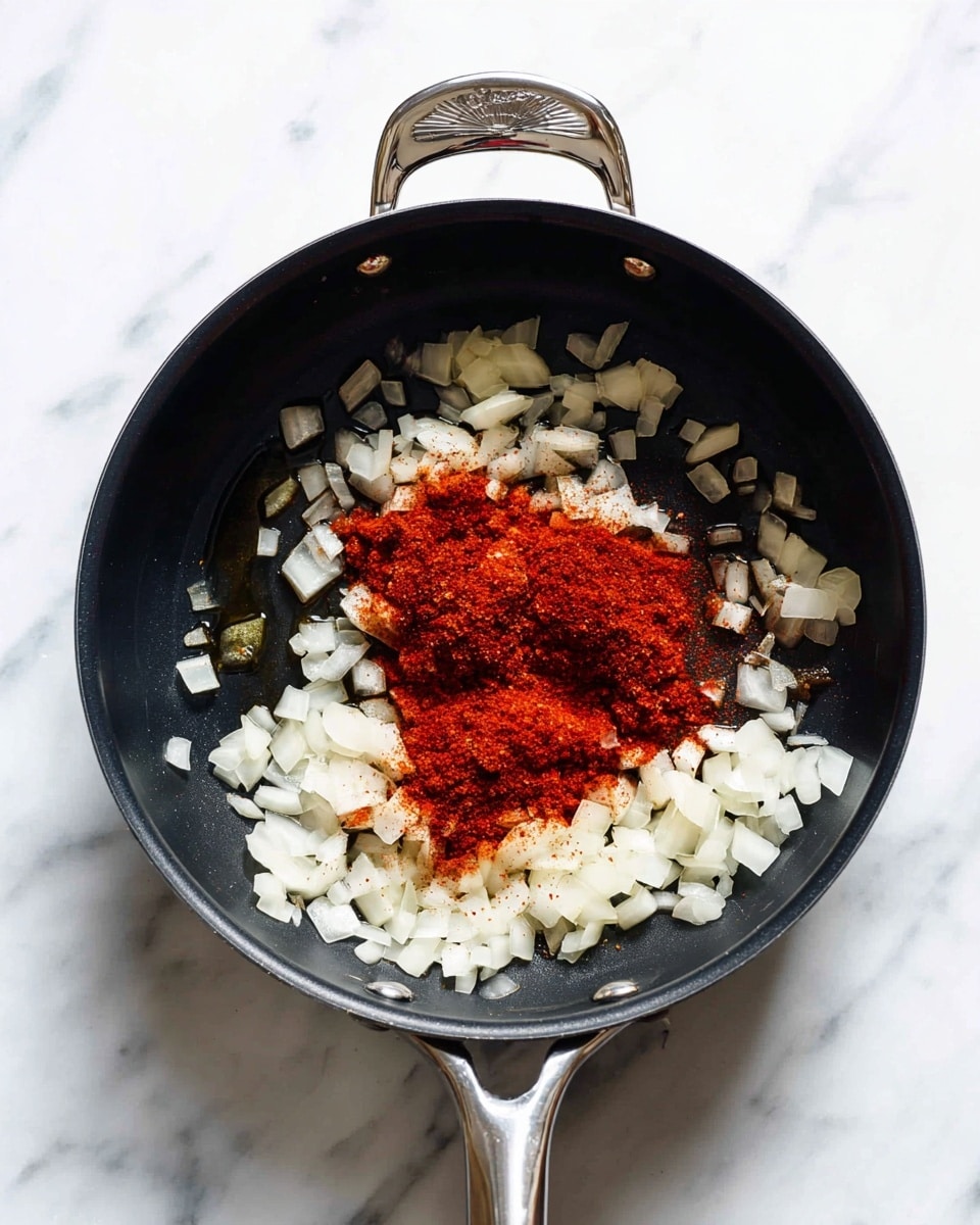 A black cooking pan with two silver handles sits on a white marbled surface. Inside the pan, finely chopped onions form an outer ring around two piles of ground red spices placed in the center. The onions are translucent with a slight shine, while the spices are a deep red with a powdery texture, layered on top of the onions. Some oil glistens slightly beneath the ingredients in the pan, highlighting the texture of the onions and spices. photo taken with an iphone --ar 4:5 --v 7