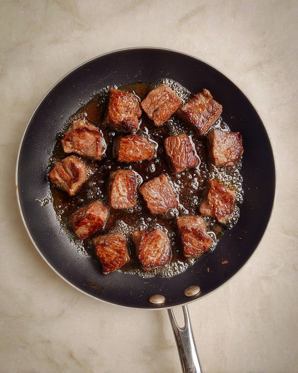A black frying pan with a silver handle holds several pieces of browned meat being cooked in bubbling oil, each piece a small cube with a seared, textured surface showing a rich brown color, all placed on a white marbled texture background. photo taken with an iphone --ar 4:5 --v 7