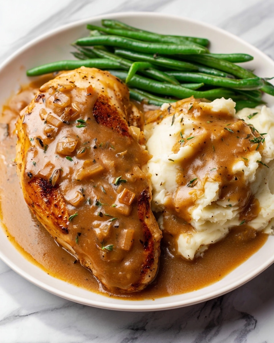 A white plate holds a meal with three main parts: a large piece of golden-brown chicken covered in thick brown gravy with small bits of onion and herbs, a creamy mound of mashed potatoes topped with the same gravy, and a small pile of green beans with a smooth, fresh texture placed next to the chicken. The gravy has a shiny, slightly textured surface with visible specks of black pepper and green herbs. The plate sits on a white marbled surface. photo taken with an iphone --ar 4:5 --v 7