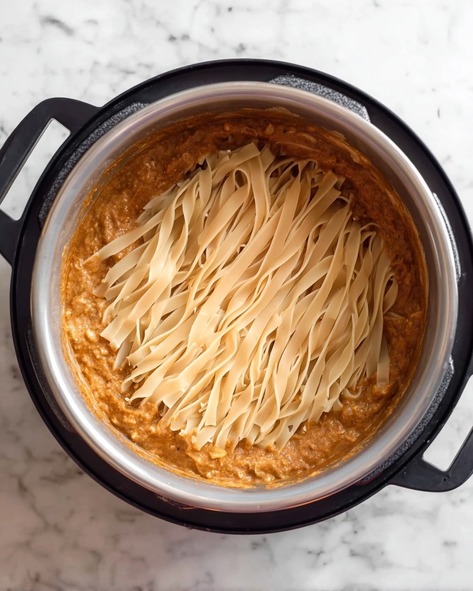 Inside a black pot on a white marbled surface, there is a dish with two layers. The bottom layer is a thick, brownish-orange sauce with a creamy texture and some visible small pieces of food. On top, there is a layer of uncooked flat pasta noodles laid out flat and covering the sauce fully, their pale beige color contrasting with the darker sauce beneath. The pot has black handles and a gray inner surface. photo taken with an iphone --ar 4:5 --v 7