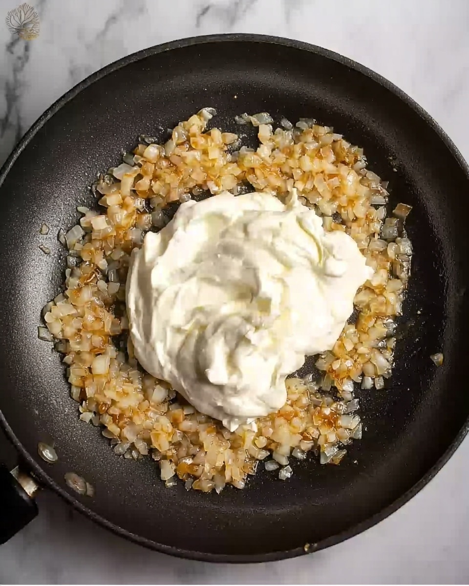The image shows a close-up view of a black frying pan on a white marbled surface, containing two main layers. The bottom layer consists of golden-brown chopped onions sautéing and spread unevenly in a ring shape around the pan. The top layer is a dollop of thick, creamy white sour cream placed in the center of the pan, surrounded on three sides by the onions. The onions have a soft, slightly oily texture, while the sour cream looks smooth and rich. Photo taken with an iphone --ar 4:5 --v 7