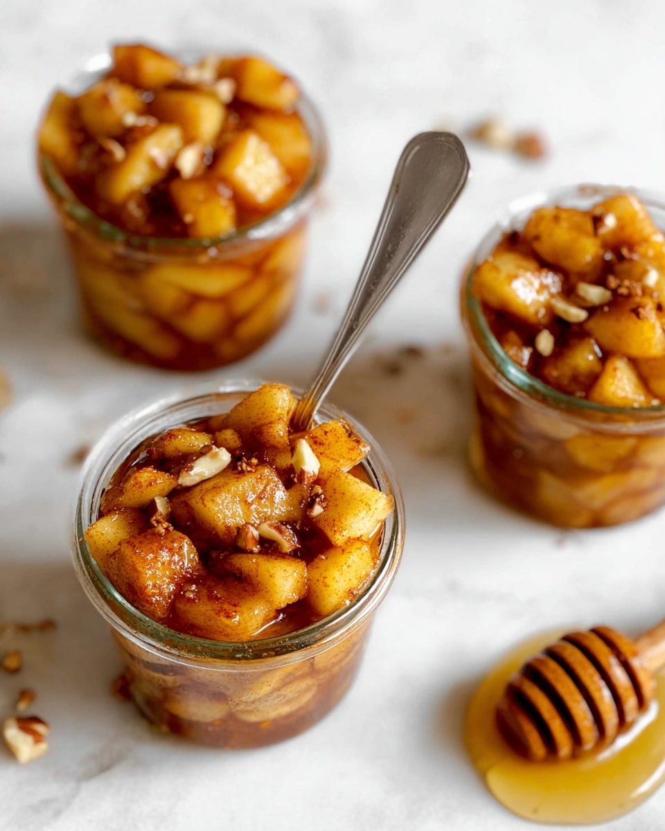 The image shows three small clear glass jars filled with a dessert topped with cooked apple chunks. Each jar has golden brown apple pieces coated in a shiny syrup with a sprinkle of cinnamon and small bits of nuts on top. One jar in the center has a silver spoon inside it, angled outwards. The jars sit on a white marbled surface that looks clean and bright. Nearby, there is a blurred honey dipper with some honey on the right side of the image. The overall look is warm and inviting with rich amber tones from the apples and syrup. photo taken with an iphone --ar 4:5 --v 7