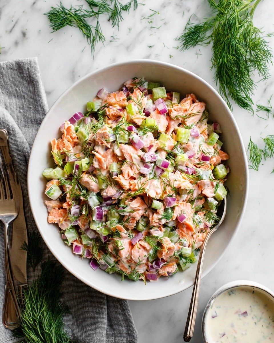 The dish is a layered salad served in a round white bowl. The first layer consists of light pink cooked salmon pieces with a soft, flaky texture. On top, there are small chunks of green celery and diced red onions, adding a crunchy texture and pops of green and purple color. Fresh chopped dill is sprinkled evenly all over, giving the dish green specks. A creamy white sauce is lightly mixed in, visible especially on the vegetables. The bowl is placed on a white marbled surface with fresh dill sprigs to the upper right, a gray cloth napkin and a fork on the left side, and a small white bowl of creamy sauce with a spoon on the lower right. The photo was taken with an iphone --ar 4:5 --v 7