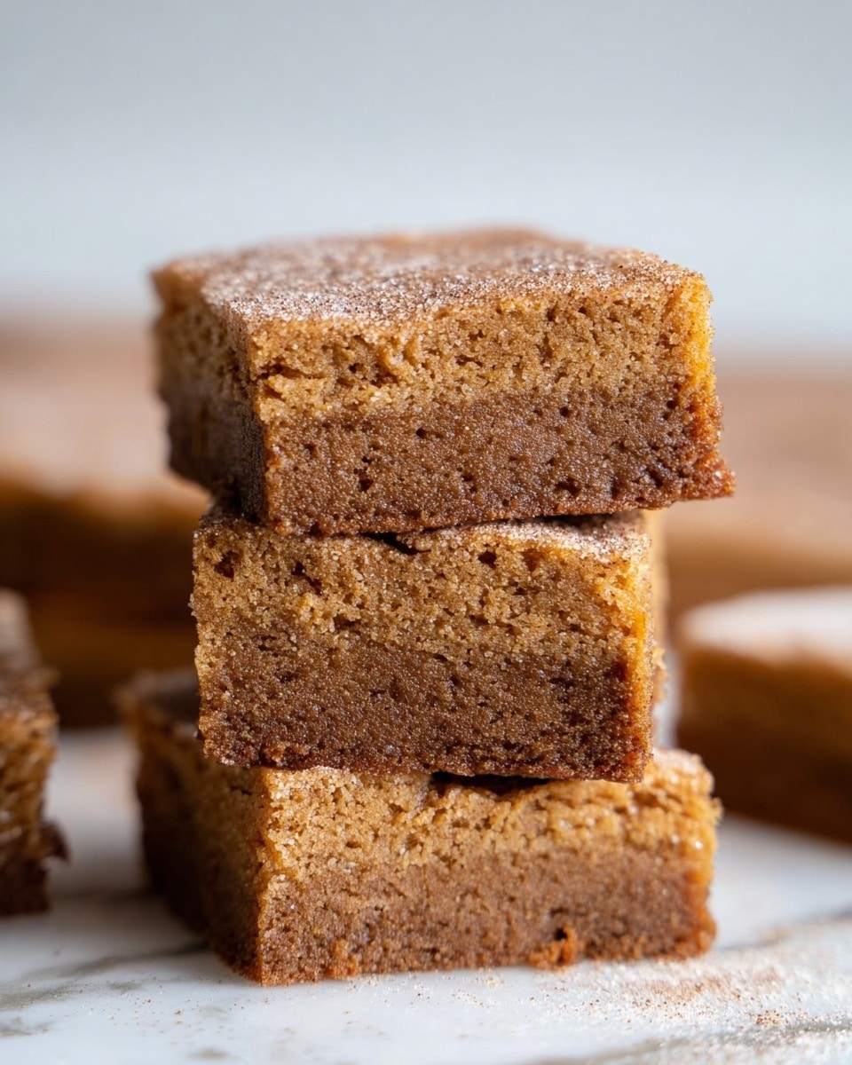 The image shows a stack of three brown cake squares on a white marbled surface with three more squares blurred in the background. Each square has one layer with a moist, dense texture and a slightly crumbly top that looks soft and spongy. The top layer has a light dusting of sugar or cinnamon, giving it a slightly grainy texture and a darker brown color than the rest of the cake. The squares are neatly cut with clear edges, and the surface they are on is smooth and clean. The focus is on the front stack, with the background softly blurred. photo taken with an iphone --ar 4:5 --v 7