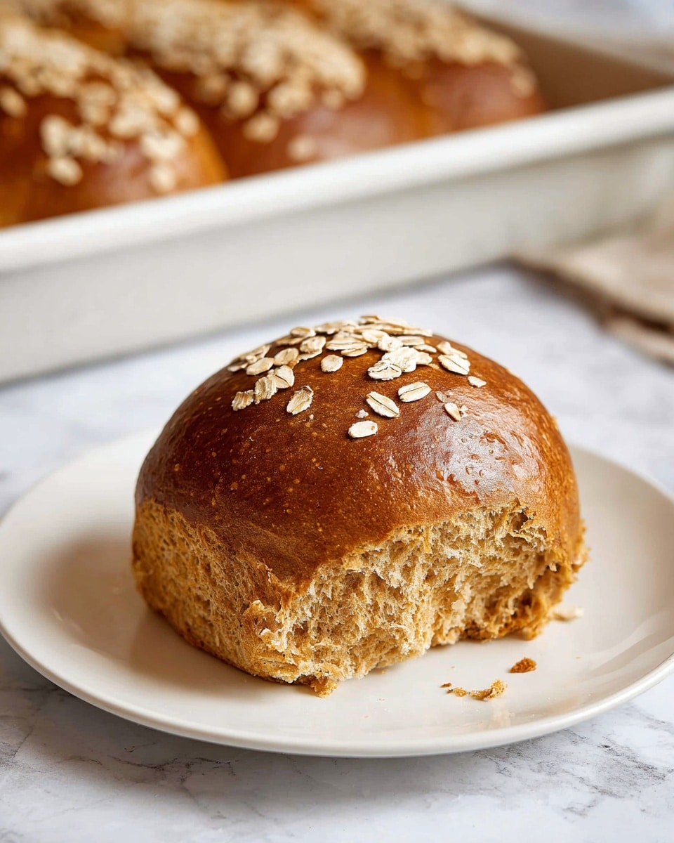 A single round brown bread roll sits on a white plate with a soft tear on one side showing its light, fluffy texture inside. The top of the roll is sprinkled with small, pale oat flakes adding a rough contrast to the smooth, shiny crust. In the background, a loaf of similar brown bread with oat flakes on top is visible inside a white baking tray, all set on a white marbled surface. Photo taken with an iphone --ar 4:5 --v 7